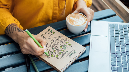 person writing in a notebook with a green pen, a cup of latte art coffee, and a laptop on a wooden table, showcasing a creative workspace.