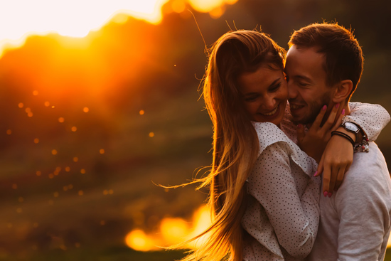 A couple joyfully embracing during sunset, with warm light illuminating their faces and a picturesque landscape in the background.