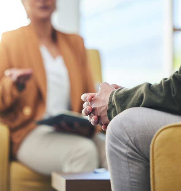 A person sitting in a yellow armchair, engaged in a conversation, with one hand gesturing while another person is seated across from them with hands clasped. The setting conveys a professional or counseling atmosphere.