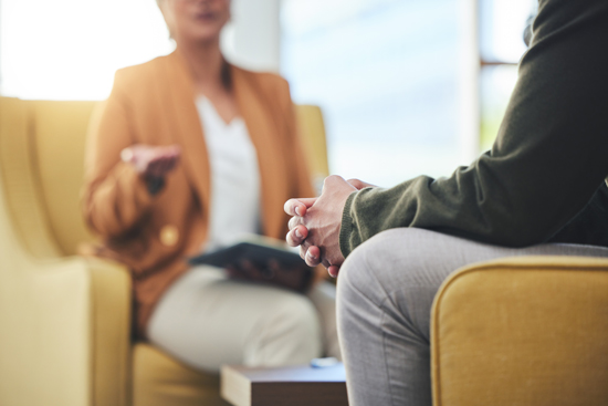 Two people engaged in a conversation, one seated in a yellow armchair while the other is partially visible, highlighting a discussion or therapy session setting.